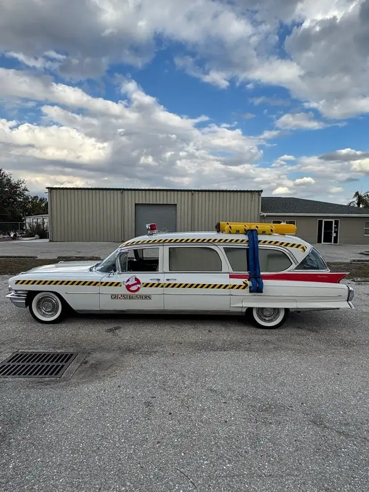 Classic white 1962 Cadillac Eureka Hearse with Ghostbusters logo and yellow-black stripes parked outside a beige industrial building, with a proton pack on its roof.