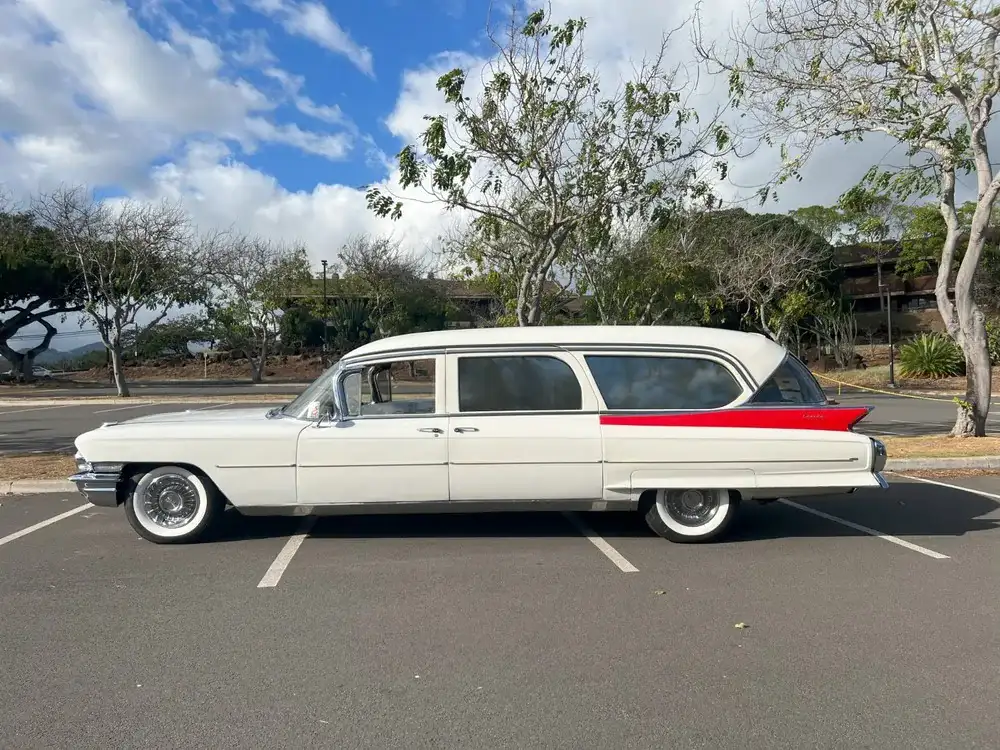 White vintage 1962 Cadillac Eureka Hearse with a red beltline parked in a sunny lot, trees in the background and a clear blue sky.