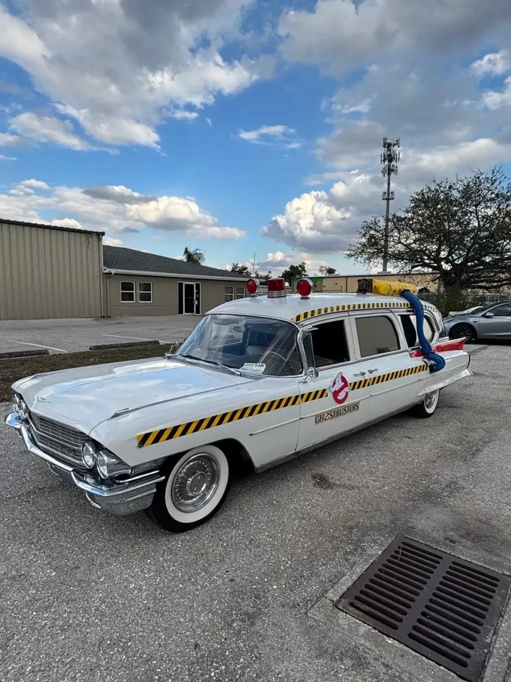 White vintage 1962 Cadillac Eureka Hearse -style Ghostbusters service car with yellow/black hazard stripes and Ghostbusters logo in a parking lot, with red emergency lights on top and a blue hose attached to the rear roof.