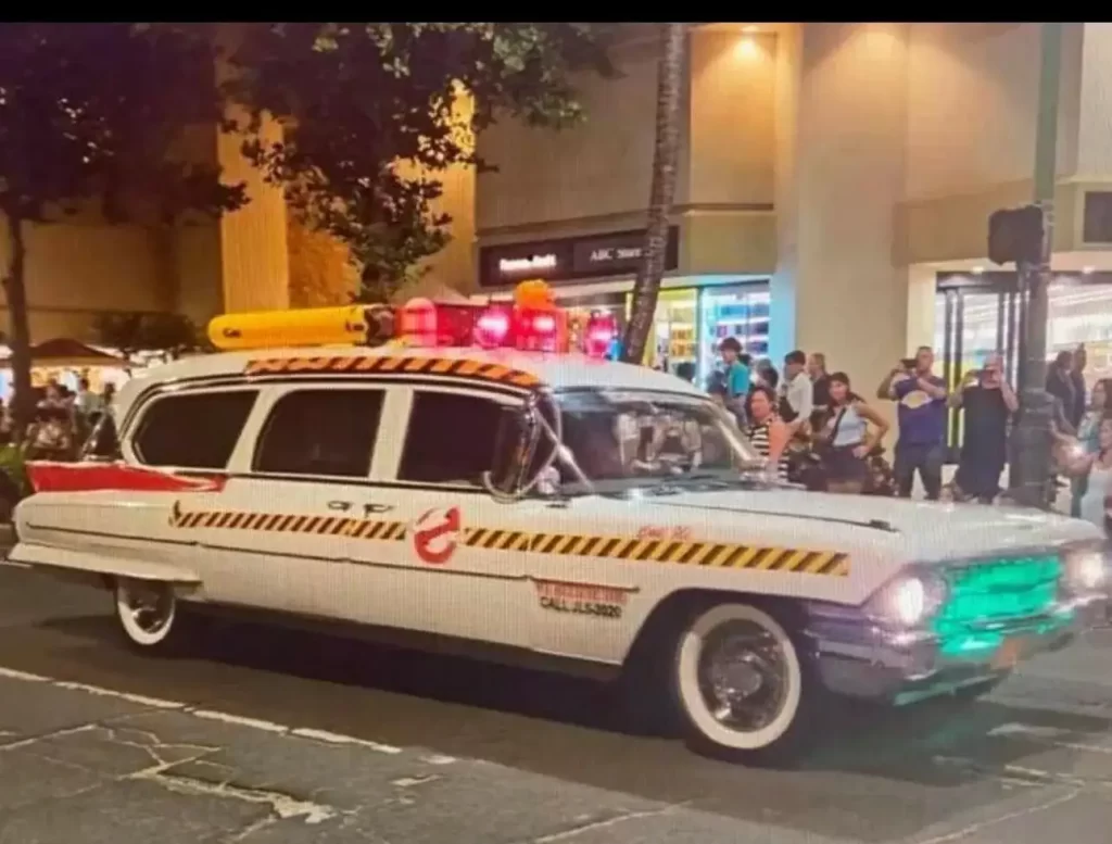 1962 Cadillac Eureka Hearse Ghostbusters Ecto-1 car with yellow-black hazard stripes on a city street at night, crowd gathered and watching.