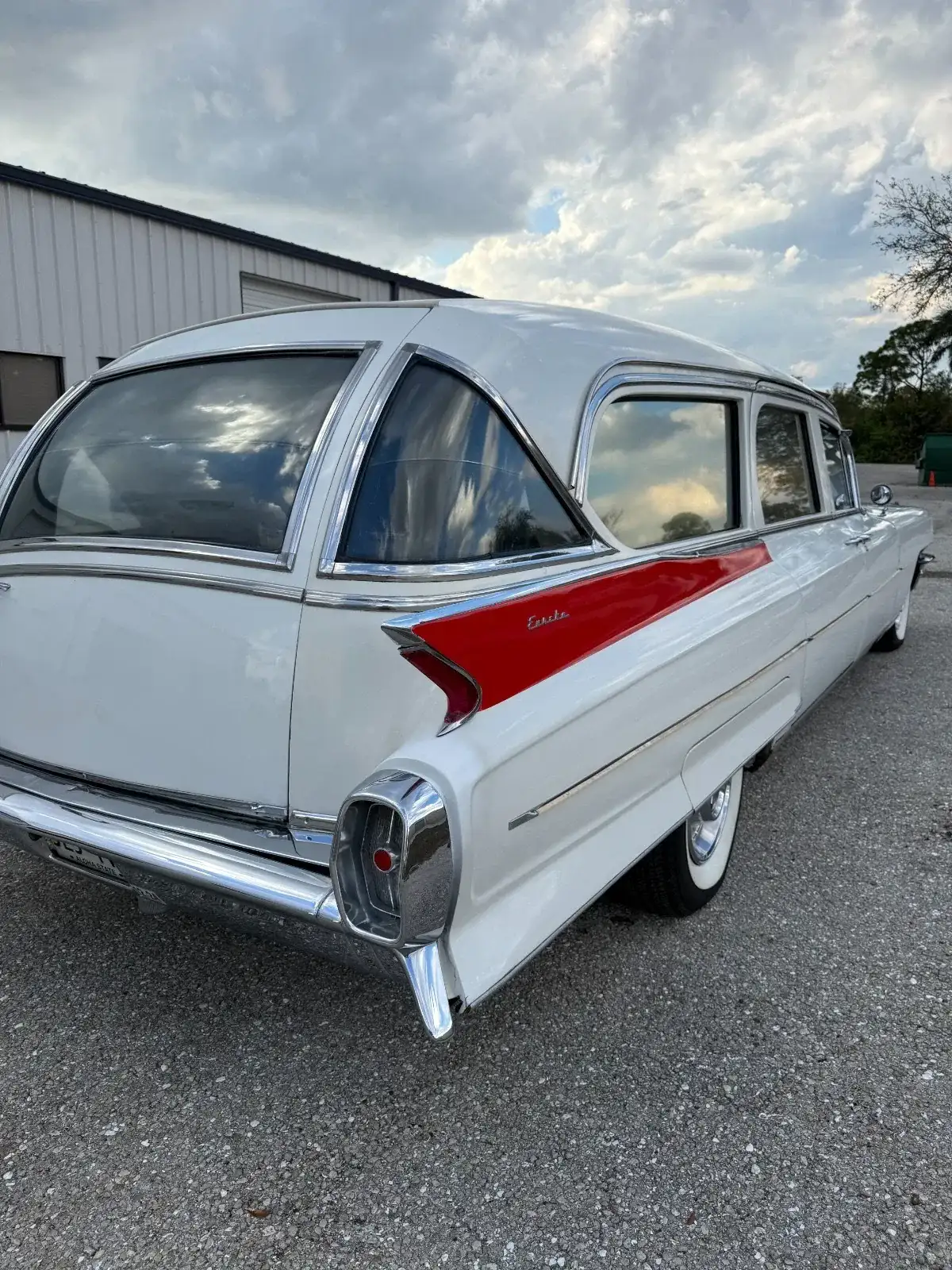 Rear three-quarter view of a white 1962 Cadillac Eureka Hearse with chrome trim, red accent on the rear quarter, and fins, parked on a lot.