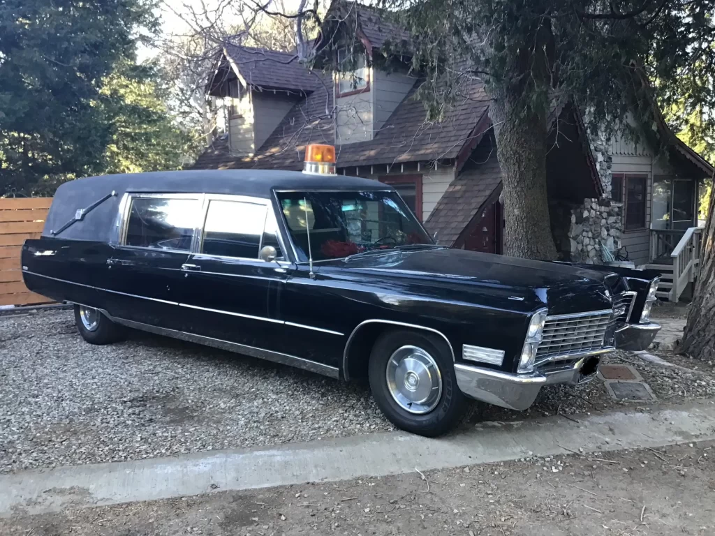 Black 1967 vintage Cadillac hearse with an orange rooftop light parked on a gravel driveway in front of a wooden house surrounded by trees.