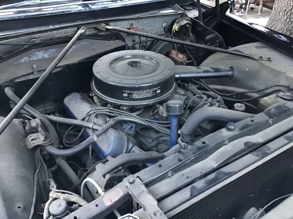 Front of a 1967 Cadillac hearse’s engine bay with a round air filter housing and dusty components visible under the hood