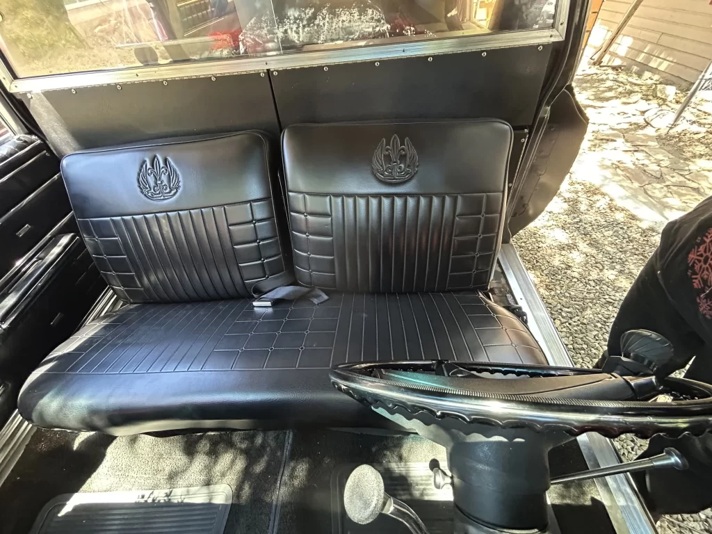 Black leather car seats with stitched pattern and a crest emblem on the headrests inside a vintage vehicle