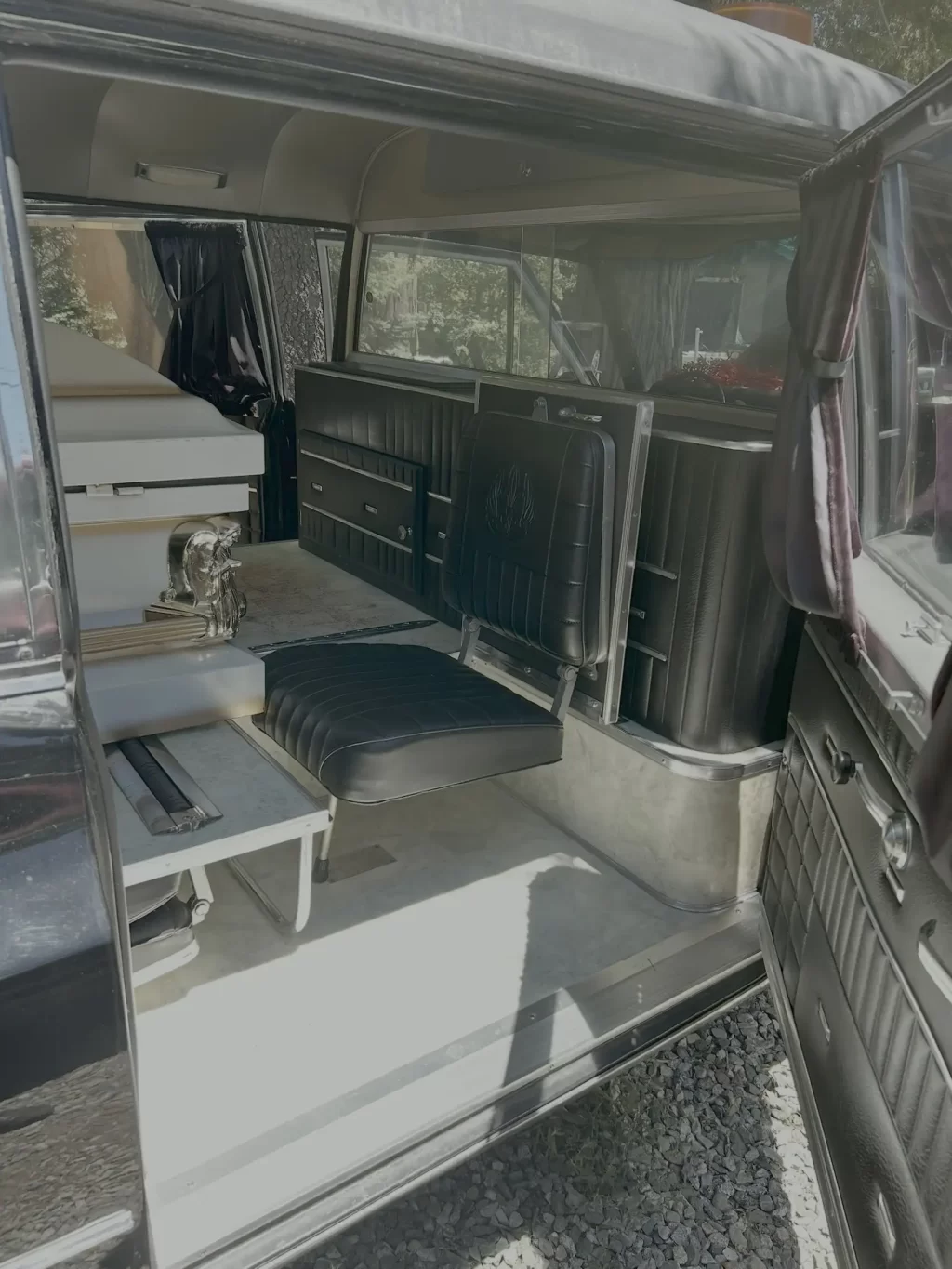 Interior of a 1967 Cadillac hearse with black jump seats and built-in storage along the side, and curtained windows.