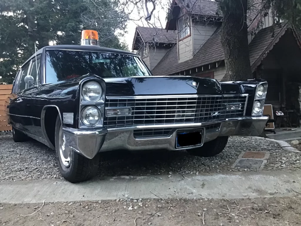 Vintage black Cadillac-style car with an orange rooftop beacon, parked in a gravel driveway in front of a wooden house with trees overhead.