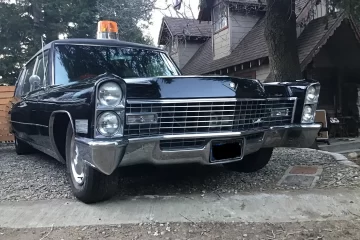 Vintage black Cadillac-style car with an orange rooftop beacon, parked in a gravel driveway in front of a wooden house with trees overhead.