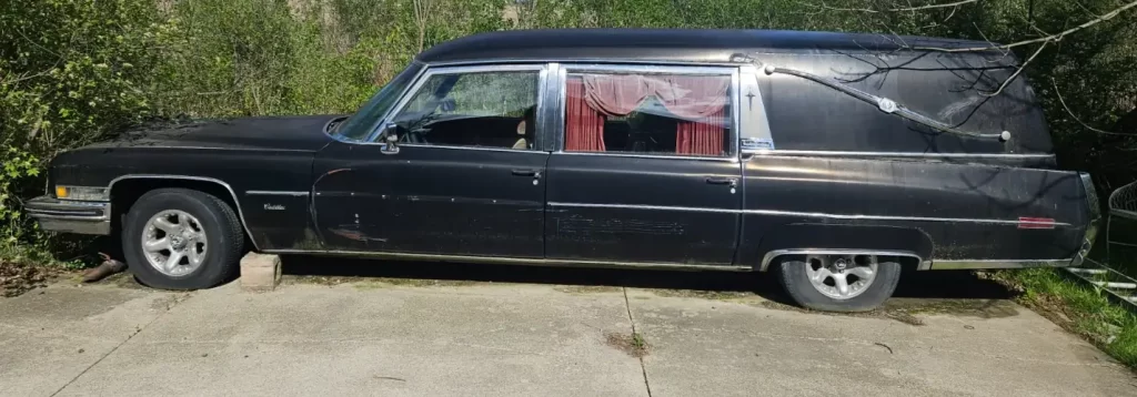 Black 1973 Cadillac Fleetwood Hearse parked on a concrete driveway, red curtains visible in the rear window.