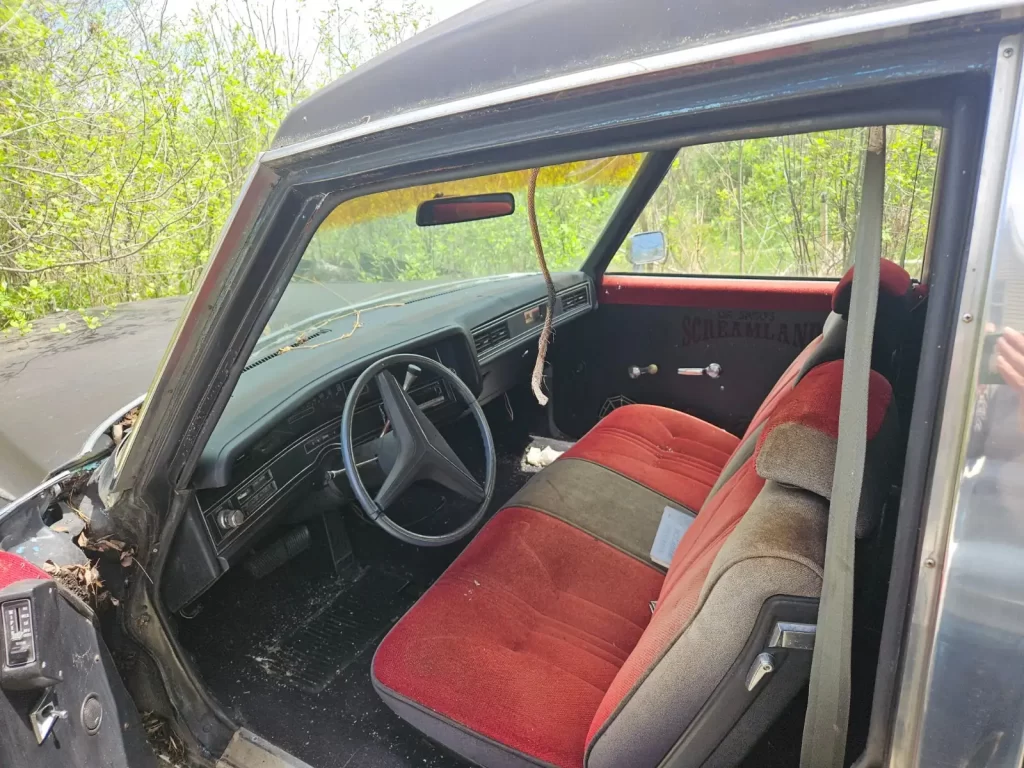 Interior of a dusty, 1973 Cadillac Fleetwood Hearse with red and tan seats, a worn dashboard, and a frayed hanging cord from the visor, surrounded by greenery outside the window.