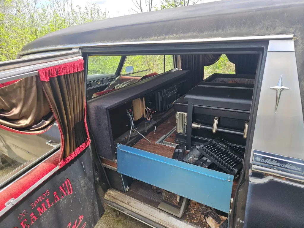 Interior of a 1973 Cadillac Fleetwood Hearse with a coffin platform and red-curtained side opening, showing black lining and chrome trim.